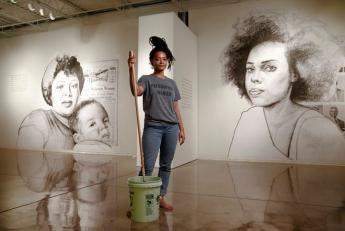 Tatyana Fazlalizadeh stans between two of her large wall paintings holding the handle of a mop inside a bucket.